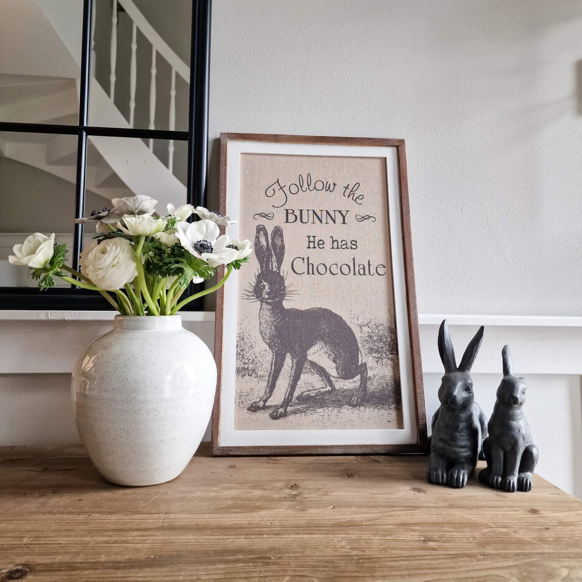 Dekoratives Bild mit Hasenmotiv und Schrift, weiße Blumen in Keramikvase, zwei Hasenfiguren auf Holztisch