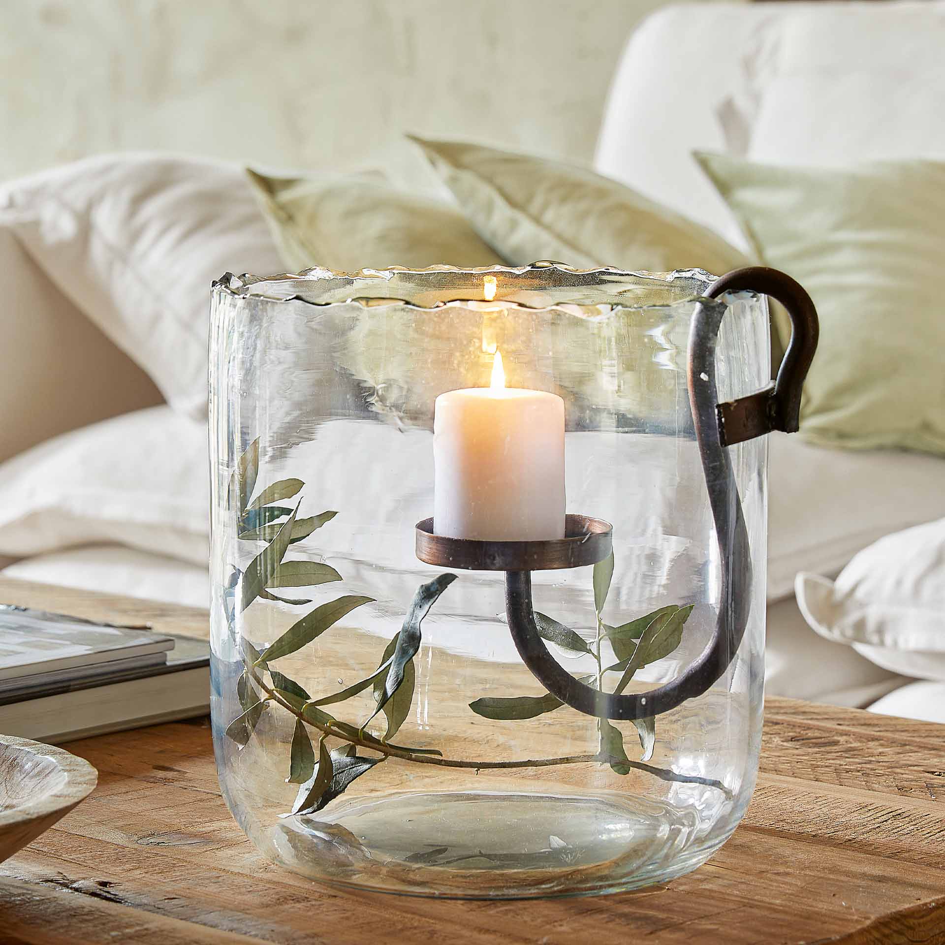 Glaswindlicht mit Metallgriff und weißer Kerze auf Holztisch mit grünen Blättern dekoriert