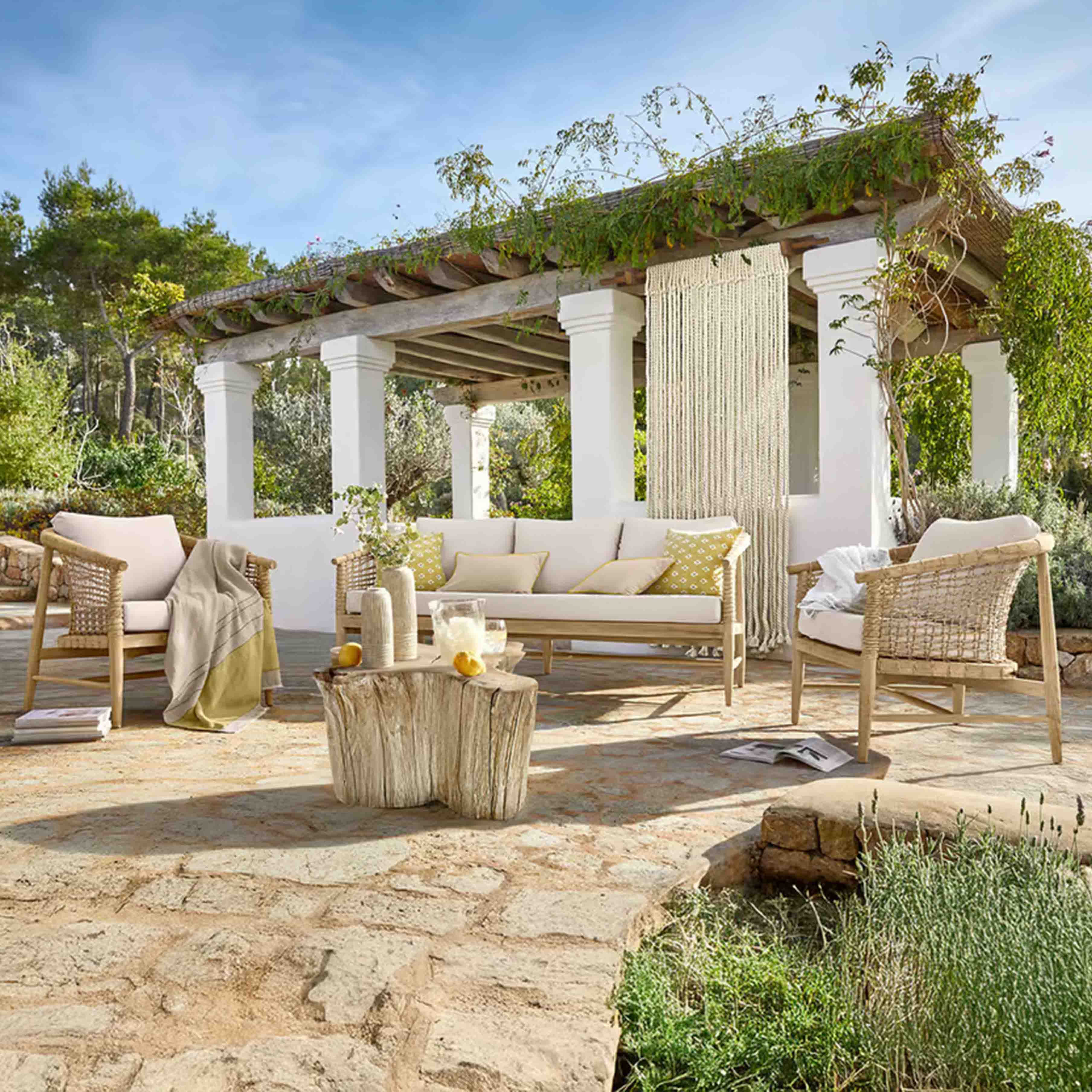 Gartenmöbel-Set aus hellem Holz mit beigen Polstern auf Steinpatio unter einer begrünten Pergola