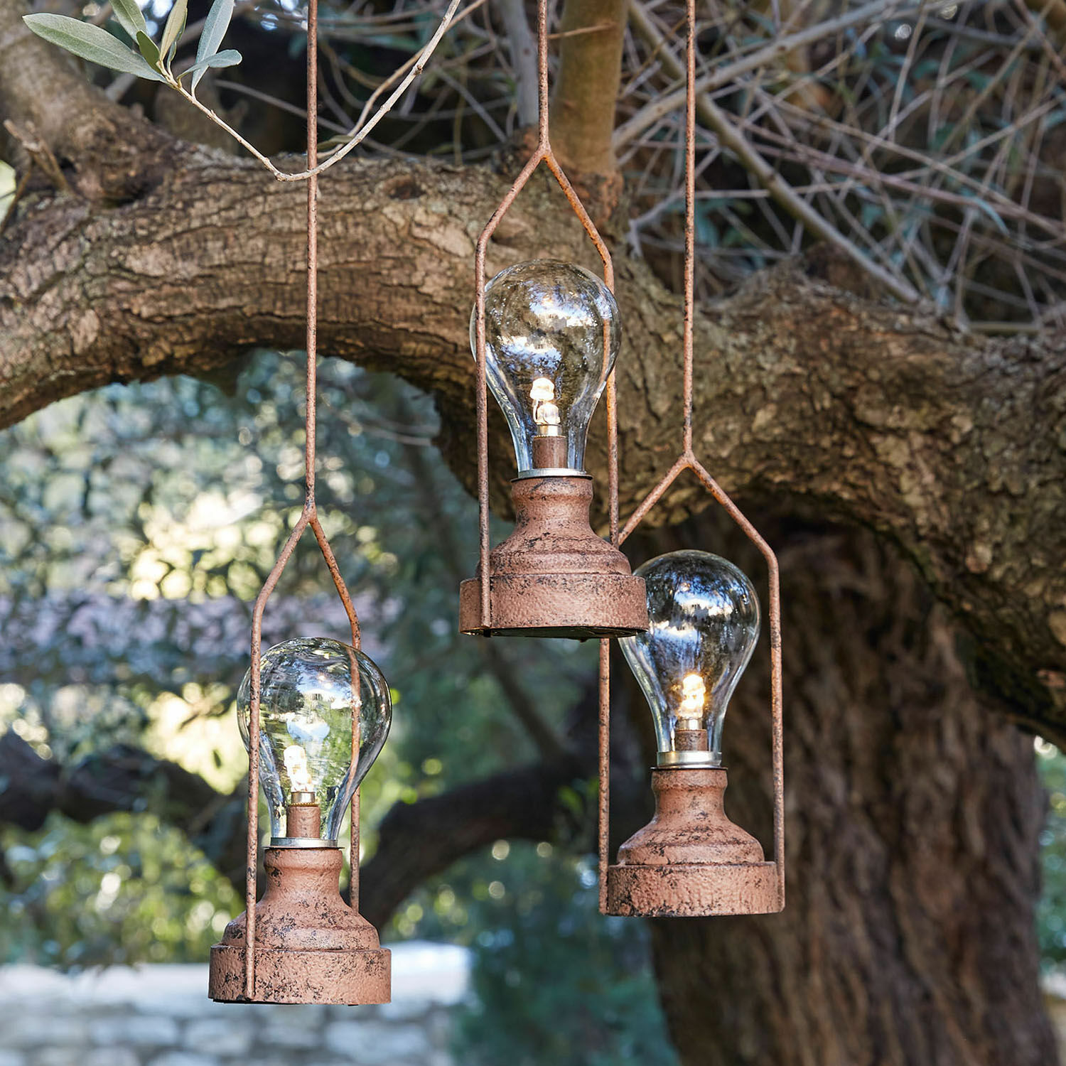 Drei hängende Lampen aus rostigem Metall mit LED-Glühbirnen vor einem Baum, dekorative Gartenbeleuchtung