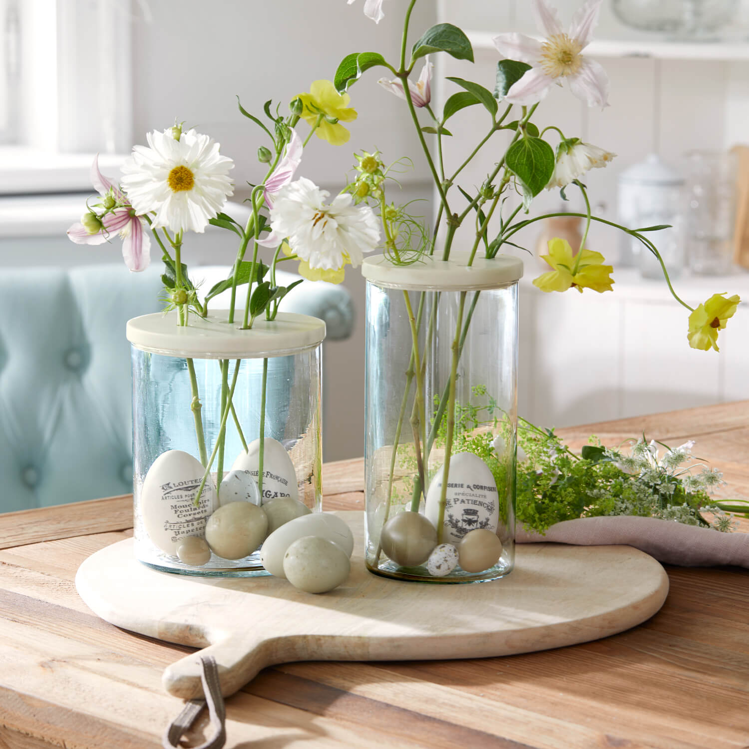 Glasvasen mit cremefarbenen Deckeln und dekorativen Eiern, pastellfarbene Blumen auf hellem Holzbrett