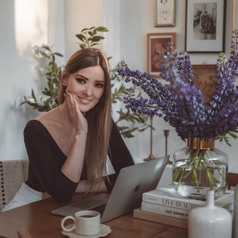 Frau arbeitet am Laptop an Holztisch mit lila Blumen in Glasvase und Kaffeetasse im gemütlichen Raum