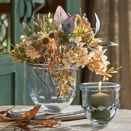 Glasvase mit getrockneten Blumen in Beige- und Brauntönen und Windlicht mit Kerze auf Holztisch