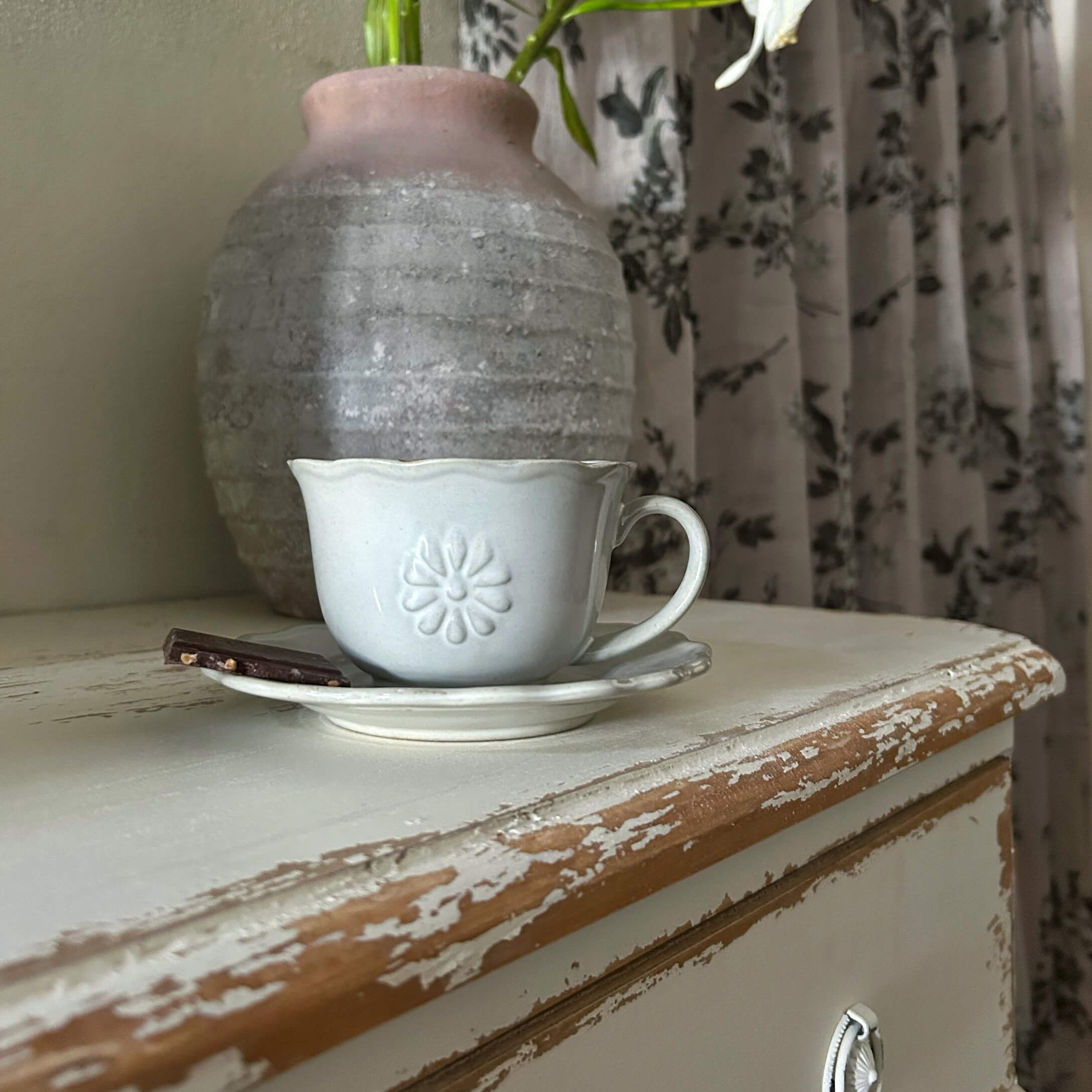Weiße Keramiktasse mit Untertasse und Schokoladenstück auf abgenutztem Holztisch