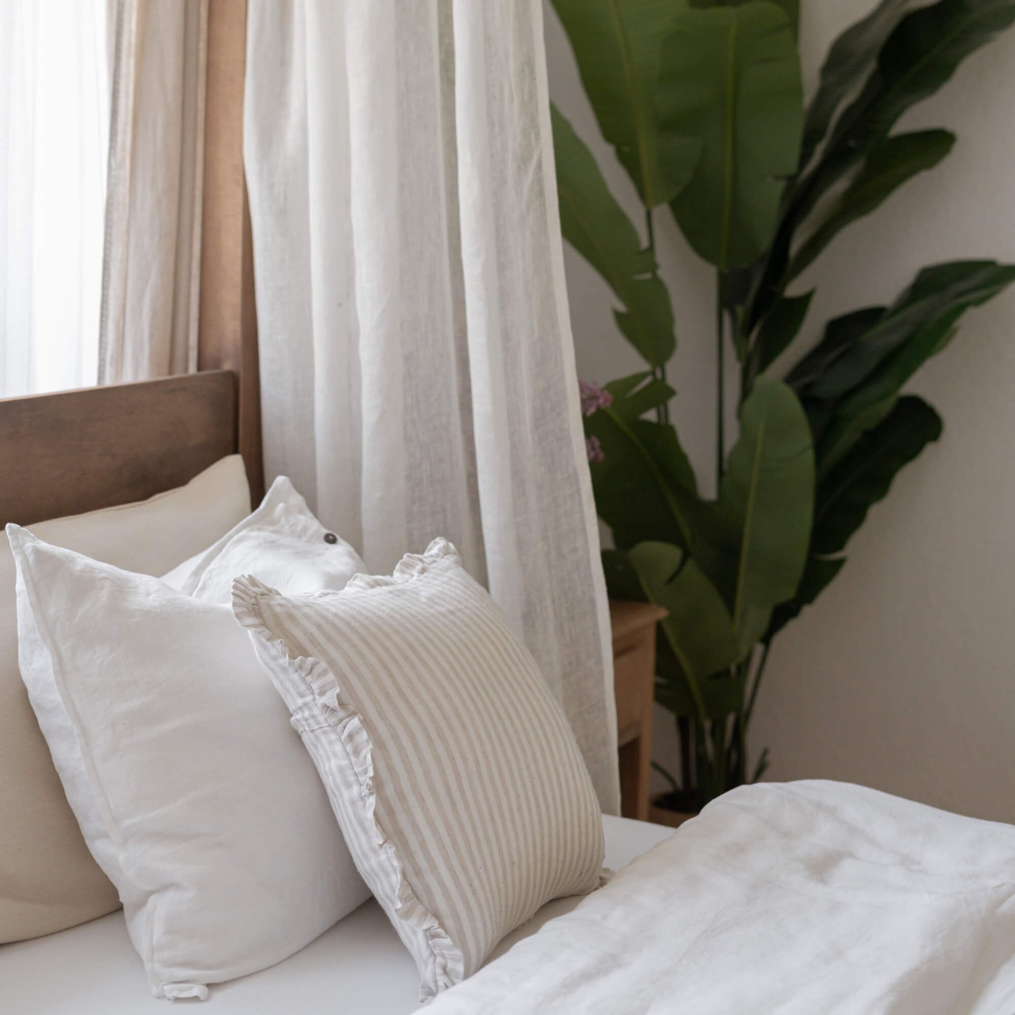 Gemütliche weiße und beige Leinenkissen auf Bett mit Holz-Kopfteil und grüner Pflanze im Hintergrund