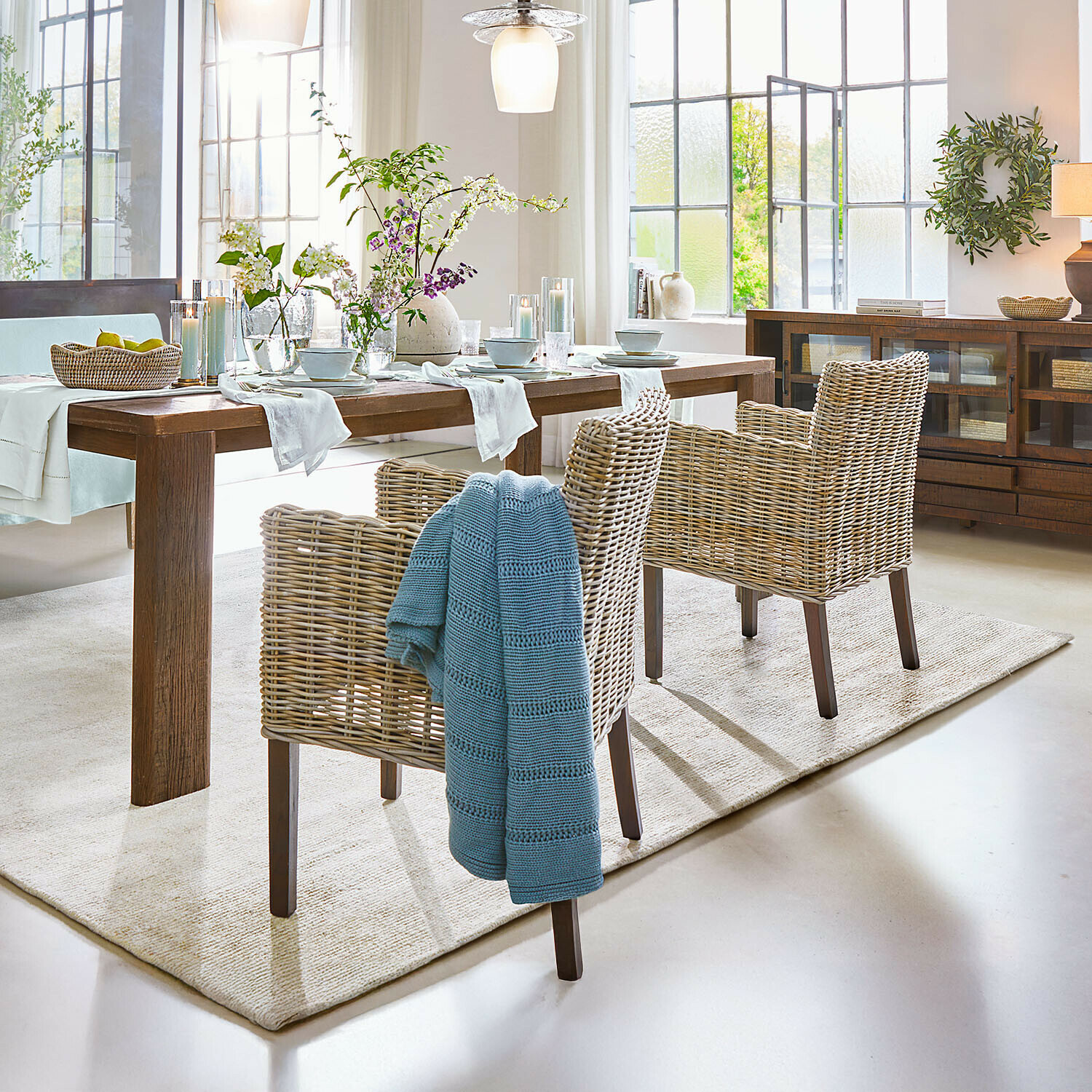 Sessel Forêt Douce Esszimmer mit Holztisch und naturfarbenen Rattanstühlen, Blumenarrangements und blauem Plaid