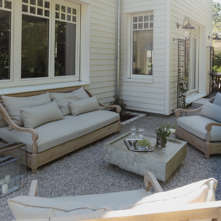 Gartenmöbel-Set aus hellem Holz mit grauen Polstern auf Kiespatio vor weißem Haus
