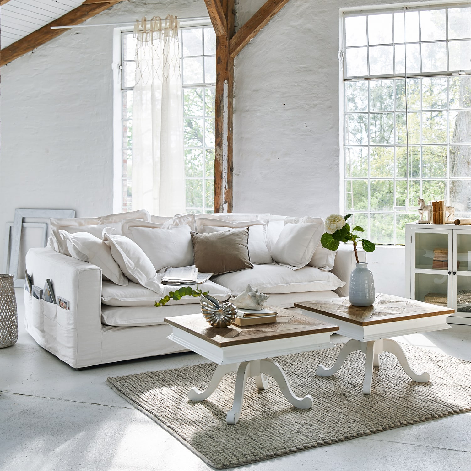Gemütliches weißes Baumwollsofa mit Kissen und zwei hölzernen Beistelltischen auf grauem Teppich
