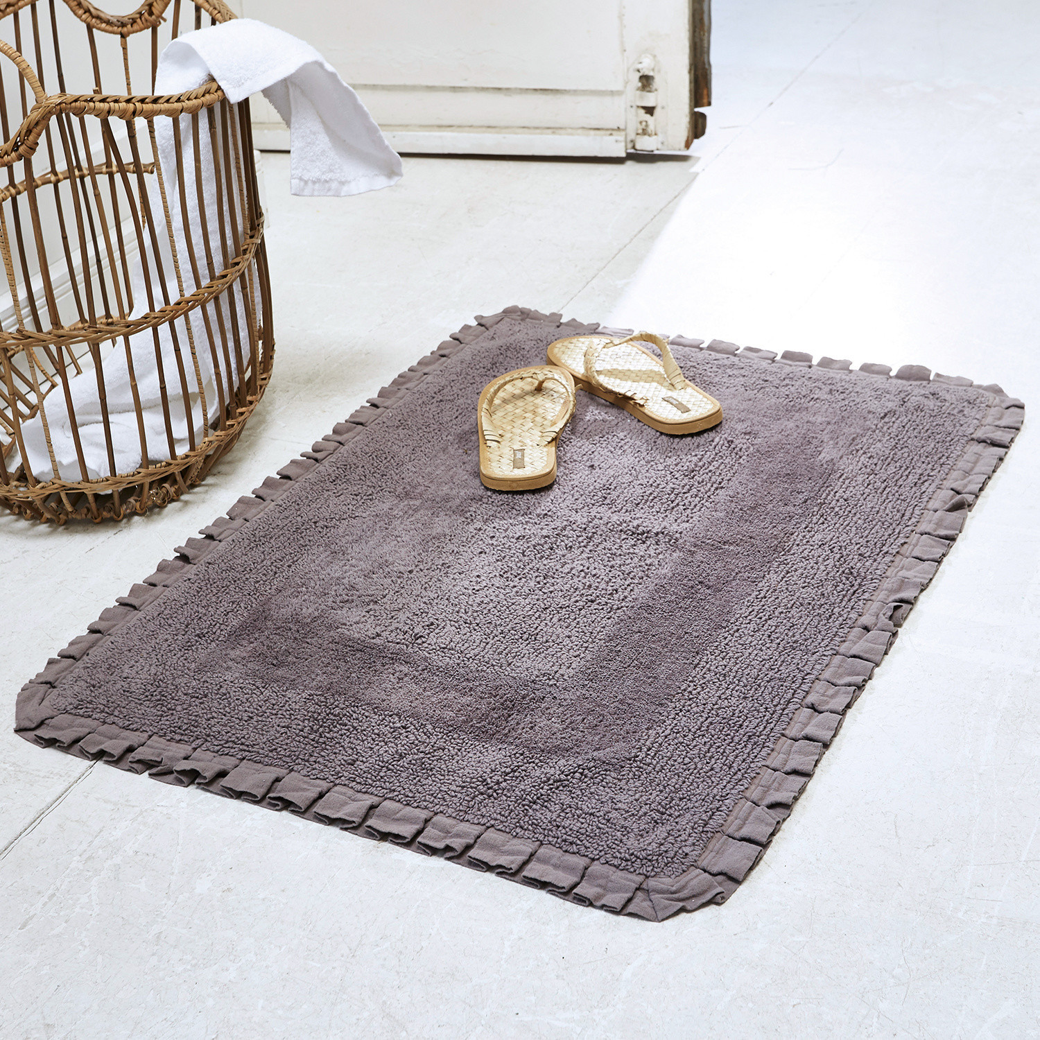 Badematte Jasmin Grauer Baumwoll-Badteppich mit Rüschensaum auf hellem Boden mit Rattansandalen