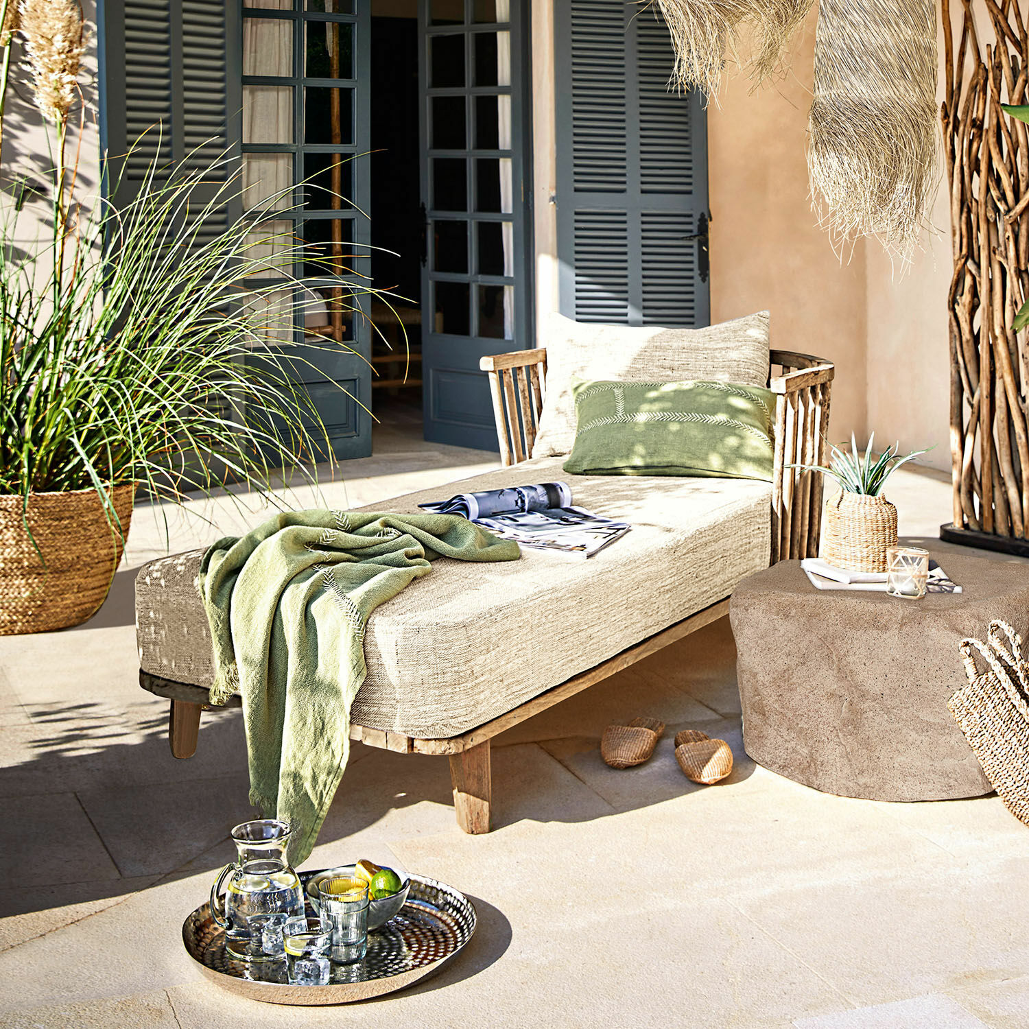 Chaiselongue Bellville Holzliege mit beigem Polster und grünem Überwurf auf Terrasse mit Dekoration und Tablett mit Getränken
