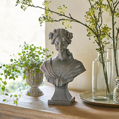 Graue Steinskulptur einer Frau mit Blumen im Haar auf Holztisch mit Pflanzenarrangement