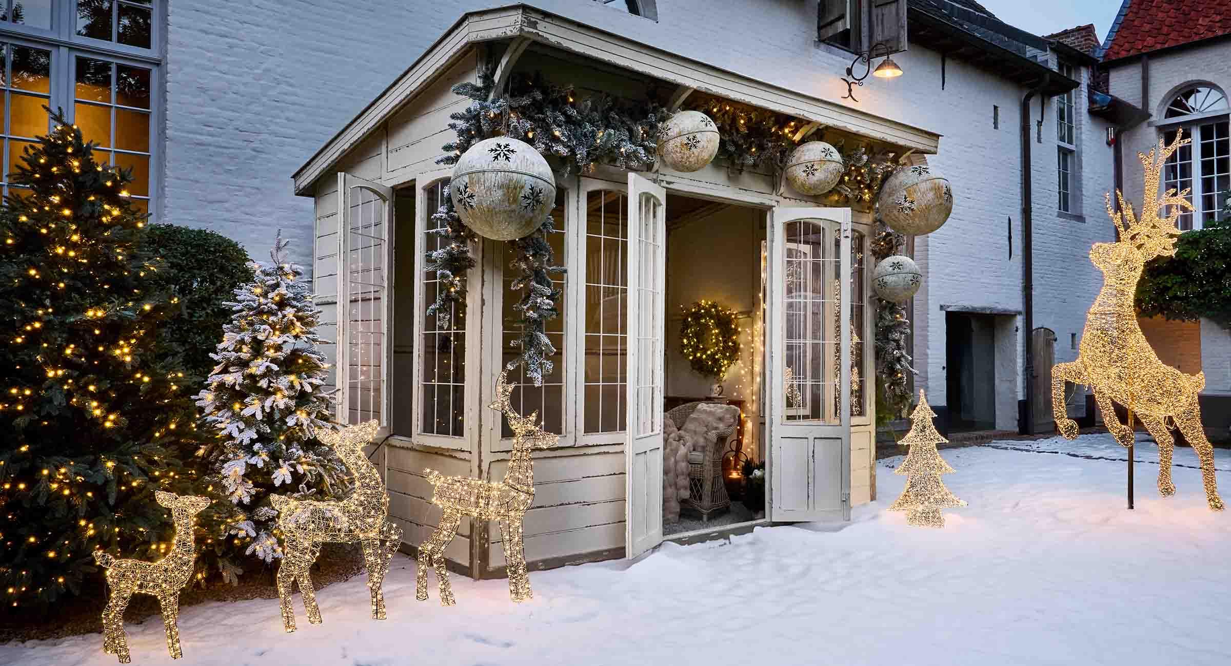 Beleuchtete Rentier- und Tannenbaum-Dekorationen im Schnee vor weißem Haus mit winterlicher Atmosphäre