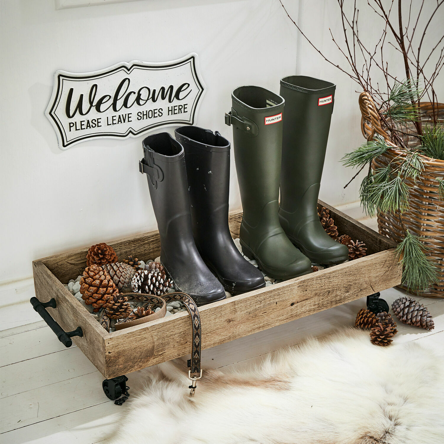 Holztablett auf Rollen mit Steinen und Kiefernzapfen, schwarze und grüne Gummistiefel im Flur