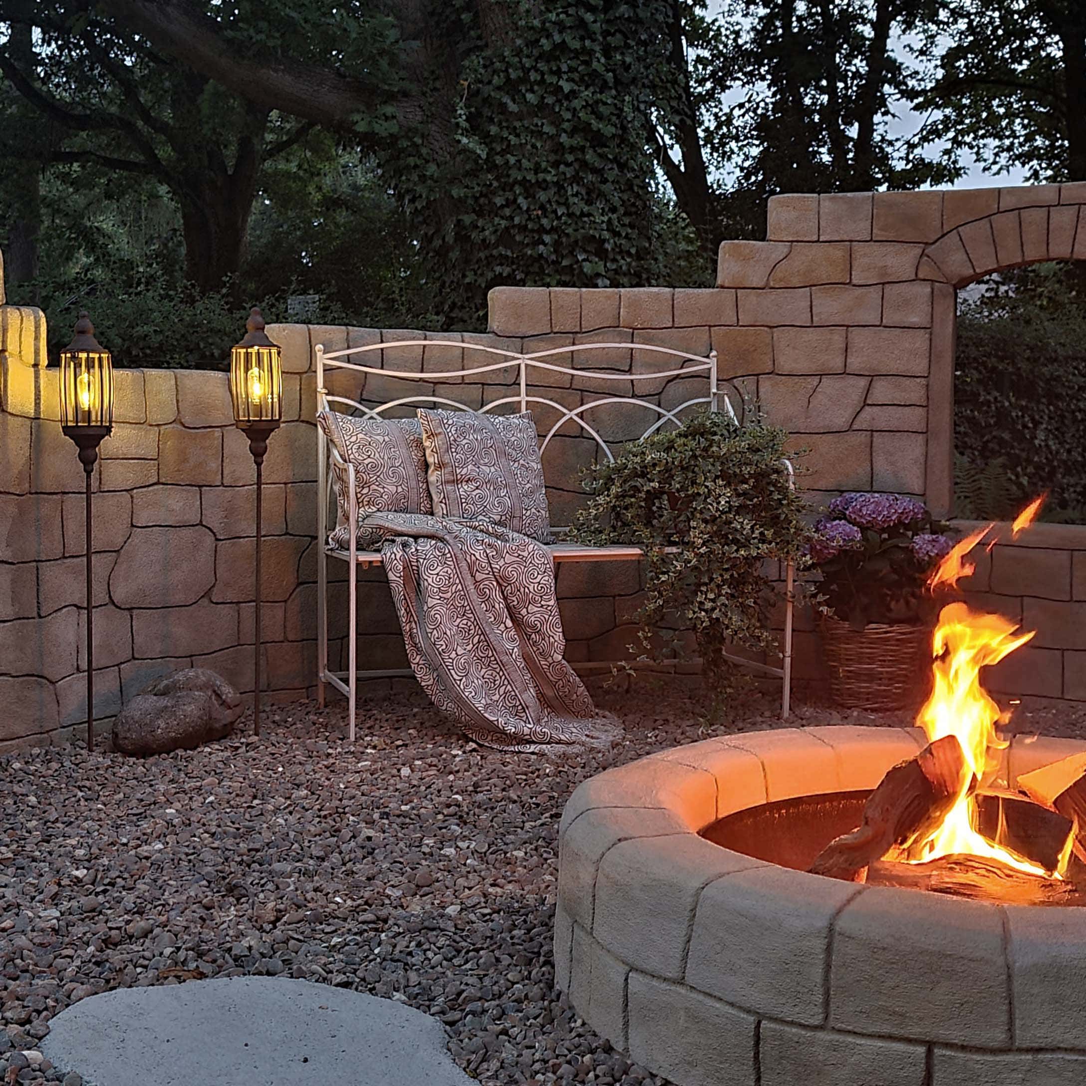 Garten mit steinernem Feuerplatz, weißer Metallbank und Stehlampen auf Kies für Außenbereich
