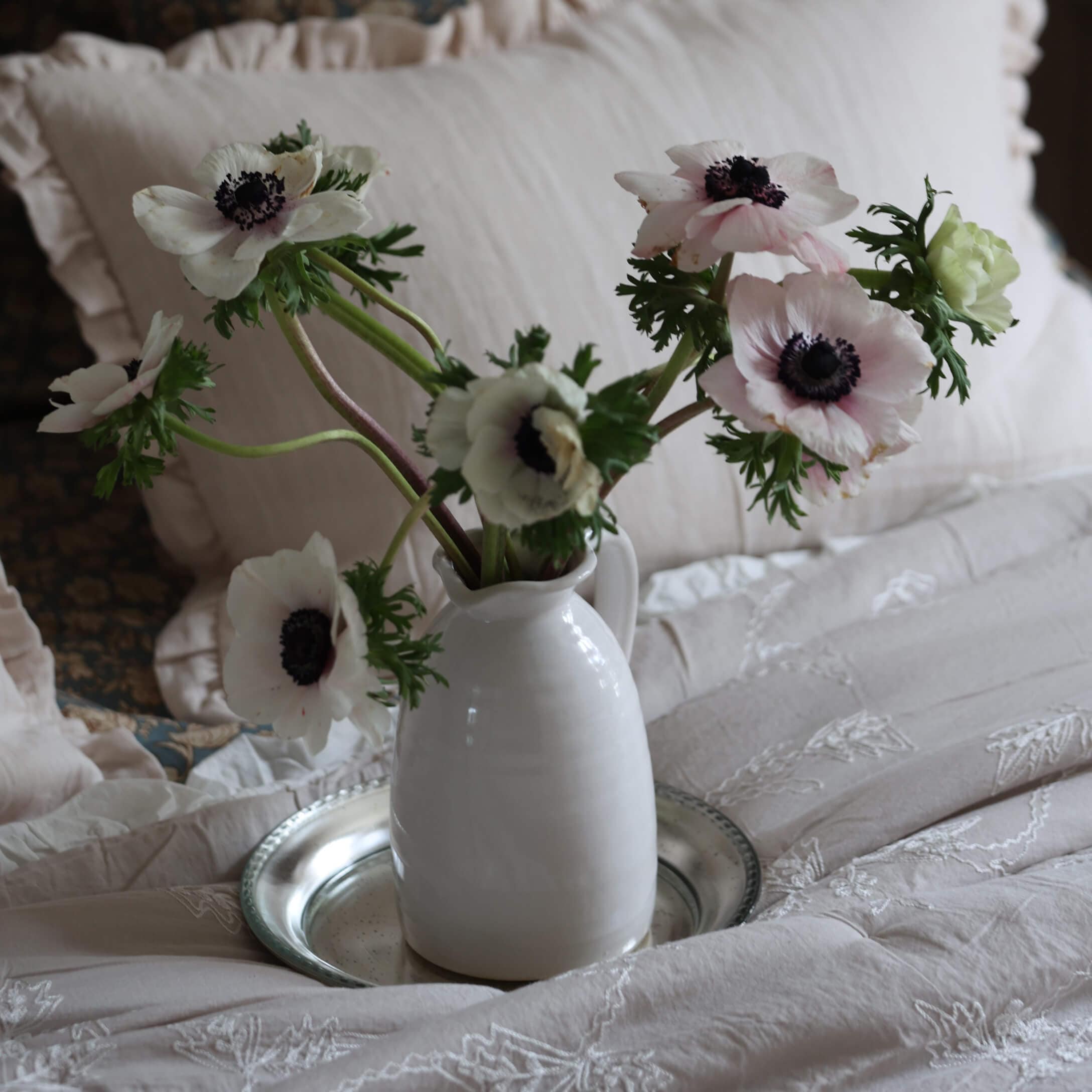 Weißer Keramikkrug mit zarten weißen Anemonen auf bestickter beiger Bettwäsche