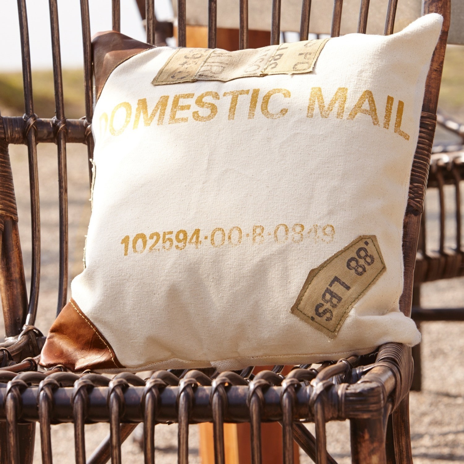 Kissen Farway Beiges Kissen mit Aufdruck Domestic Mail und braunen Leder-Ecken auf einem Rattanstuhl