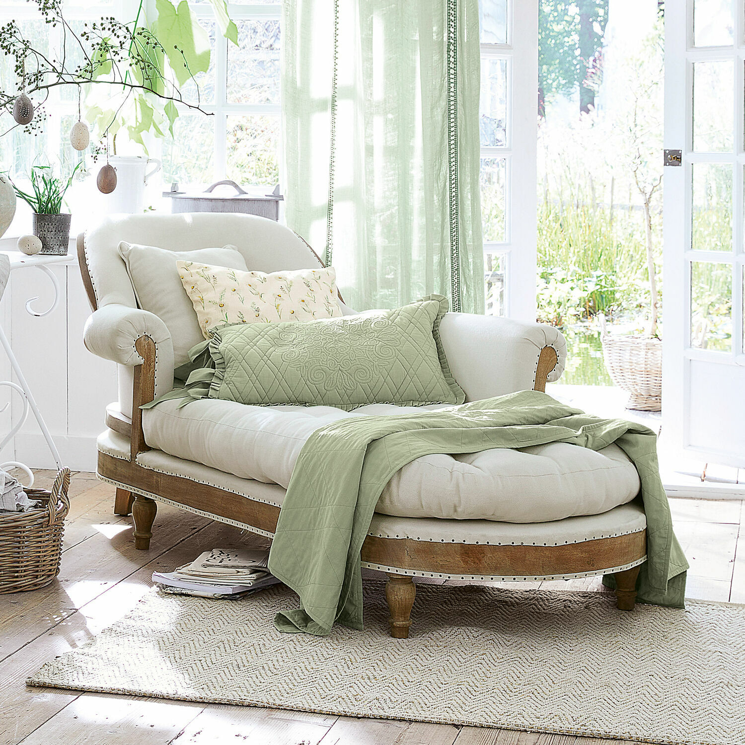 Chaiselongue Queyranne Elegante Chaiselongue aus hellem Holz und cremefarbenem Stoff mit grünem Überwurf und Kissen in hellem Raum