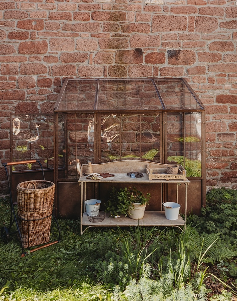 Garten-Gewächshaus aus Glas mit Metallrahmen und Holztisch vor roter Backsteinmauer