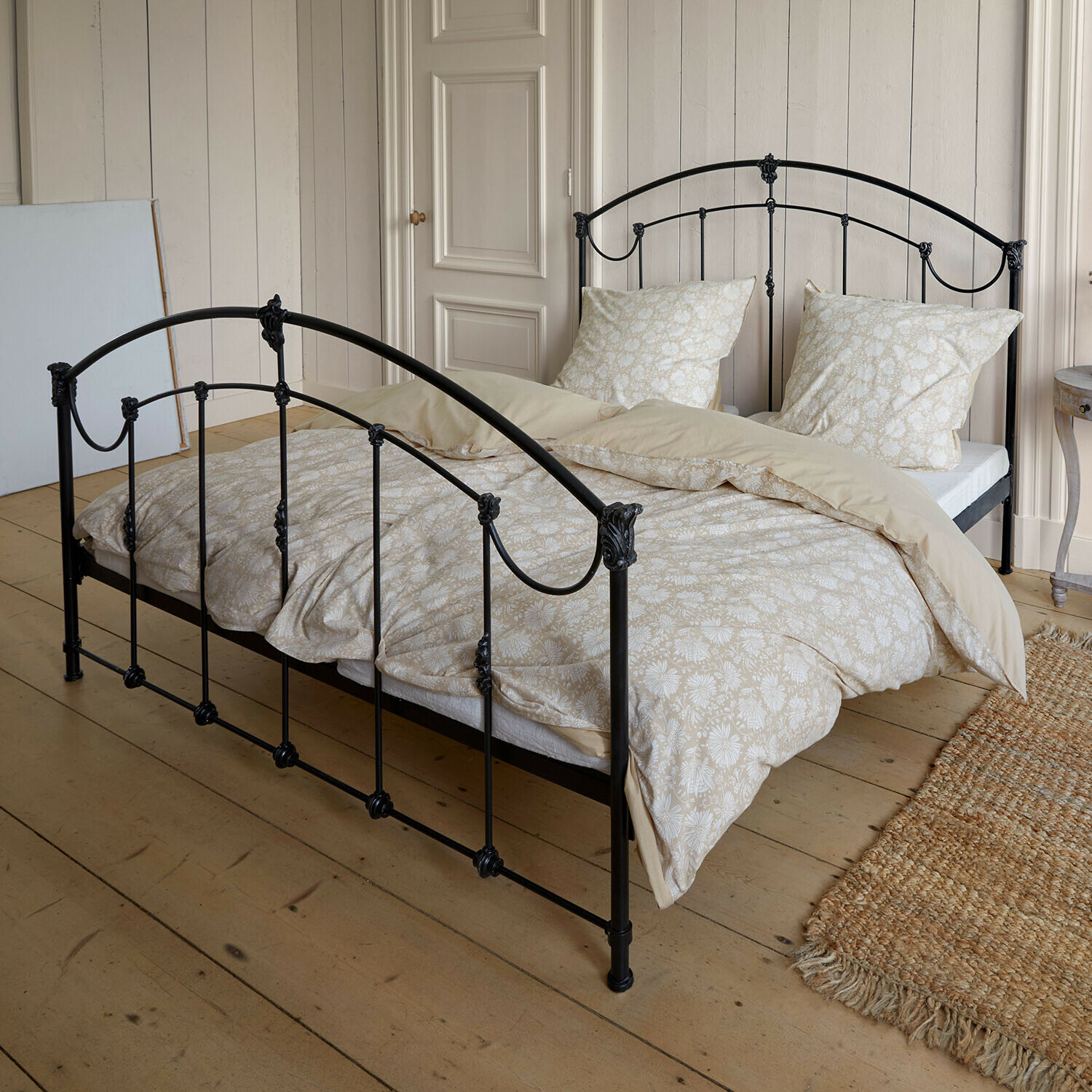 Bett Canefield Schwarzes Metallbett mit beiger Bettwäsche im Blumenmuster auf hellem Holzboden