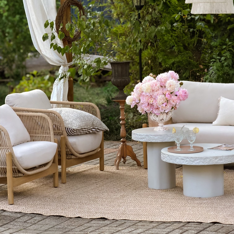 Gartenmöbel-Set mit hellen Kissen, Rattansesseln und Betontischen auf Juteteppich im Außenbereich