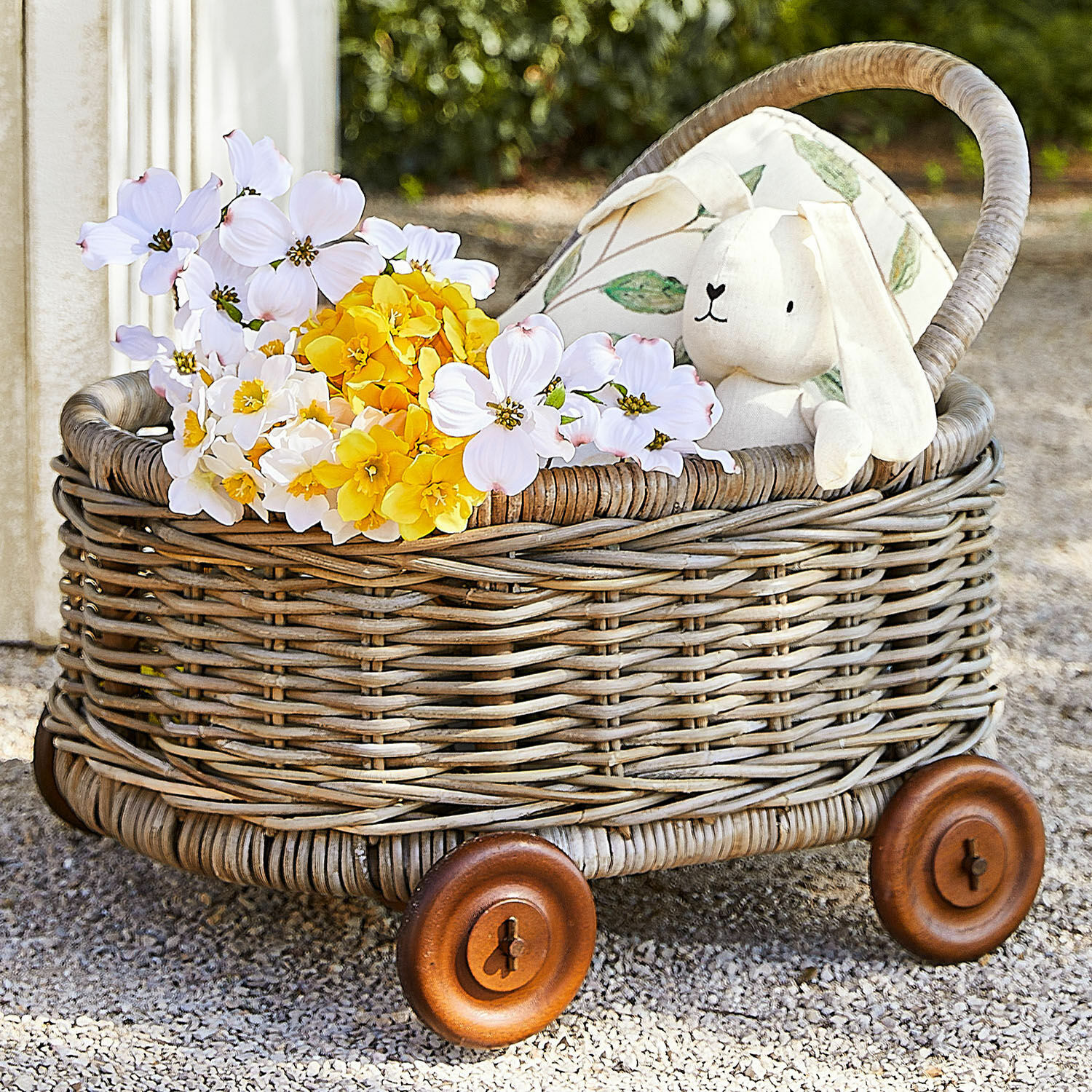 Rollwagen Narivena Wagen aus Rattan mit Holzrädern, gefüllt mit gelben und weißen Blumen und einem Plüschhasen