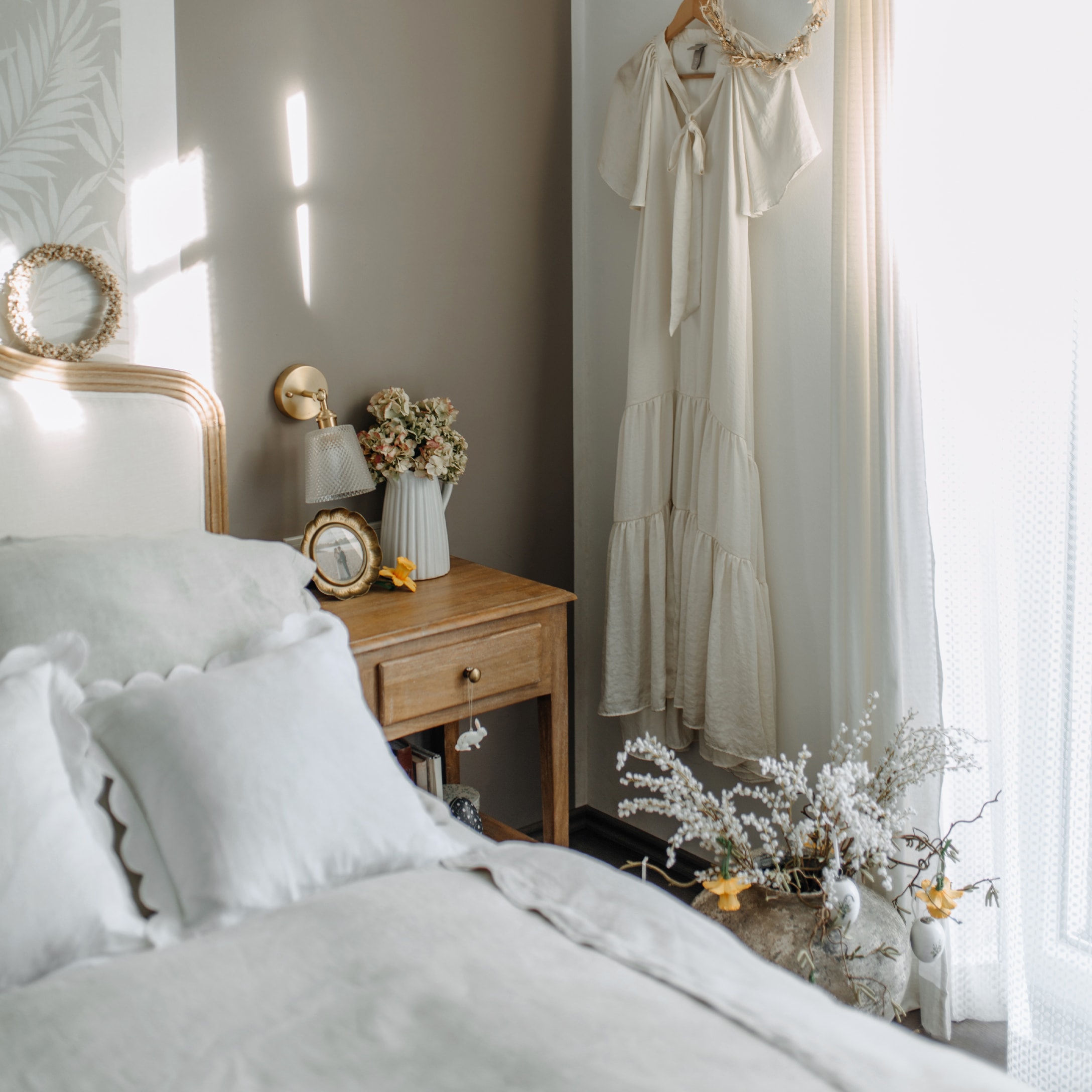 Gemütliches Schlafzimmer mit Holznachttisch, weißer Bettwäsche und cremefarbenem Kleid an der Wand