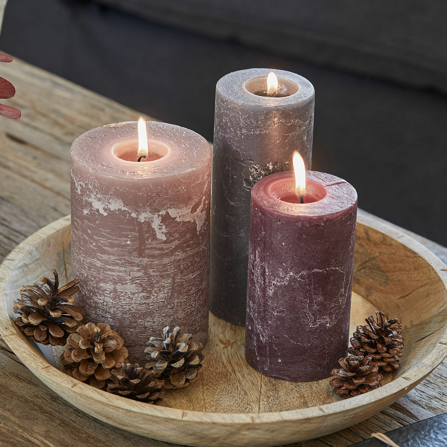 Drei brennende Kerzen in Rosa- und Grautönen auf Holztablett mit Kiefernzapfen dekoriert