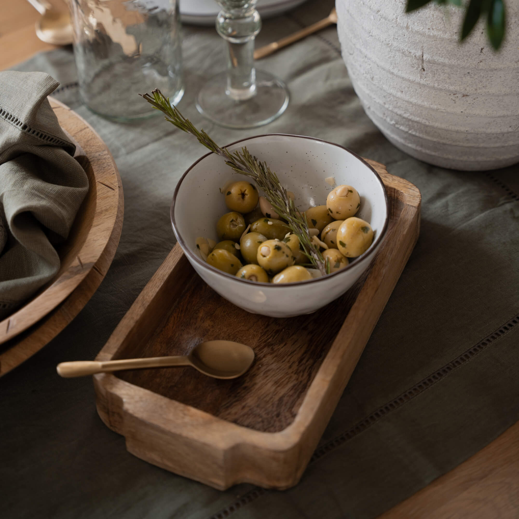 Weiße Keramikschale mit grünen Oliven und Rosmarinzweig auf Holztablett auf dem Tisch