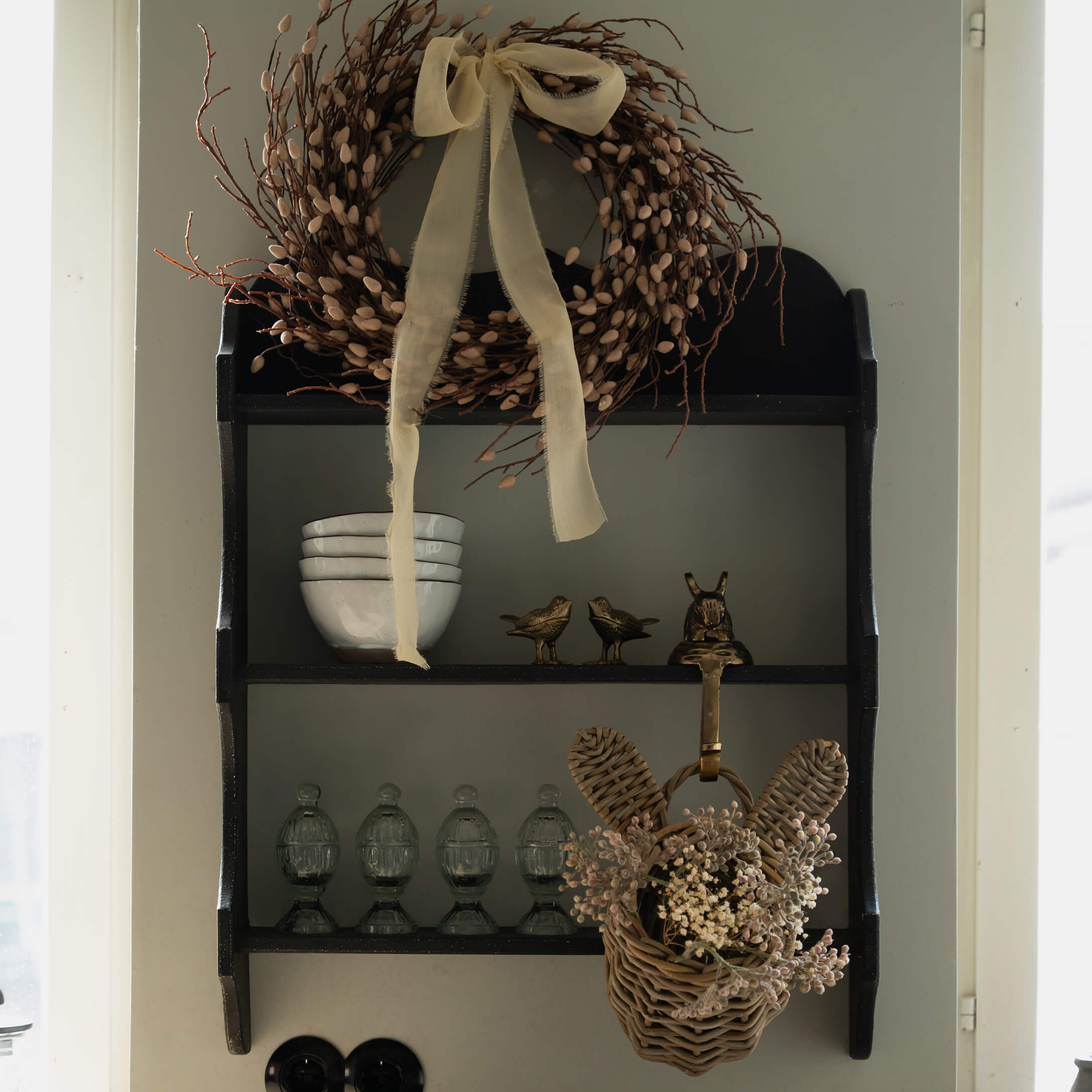 Schwarzes Holzregal mit dekorativem Kranz und Weidenkorb mit Trockenblumen an der Wand