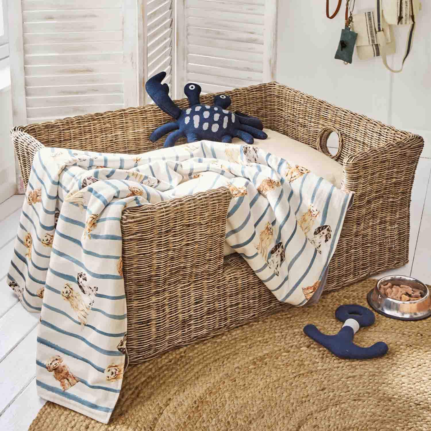 Wicker-Hundebett mit cremefarbenem Kissen, gestreifter Decke mit Hundemotiven und dunkelblauem Krabben-Spielzeug