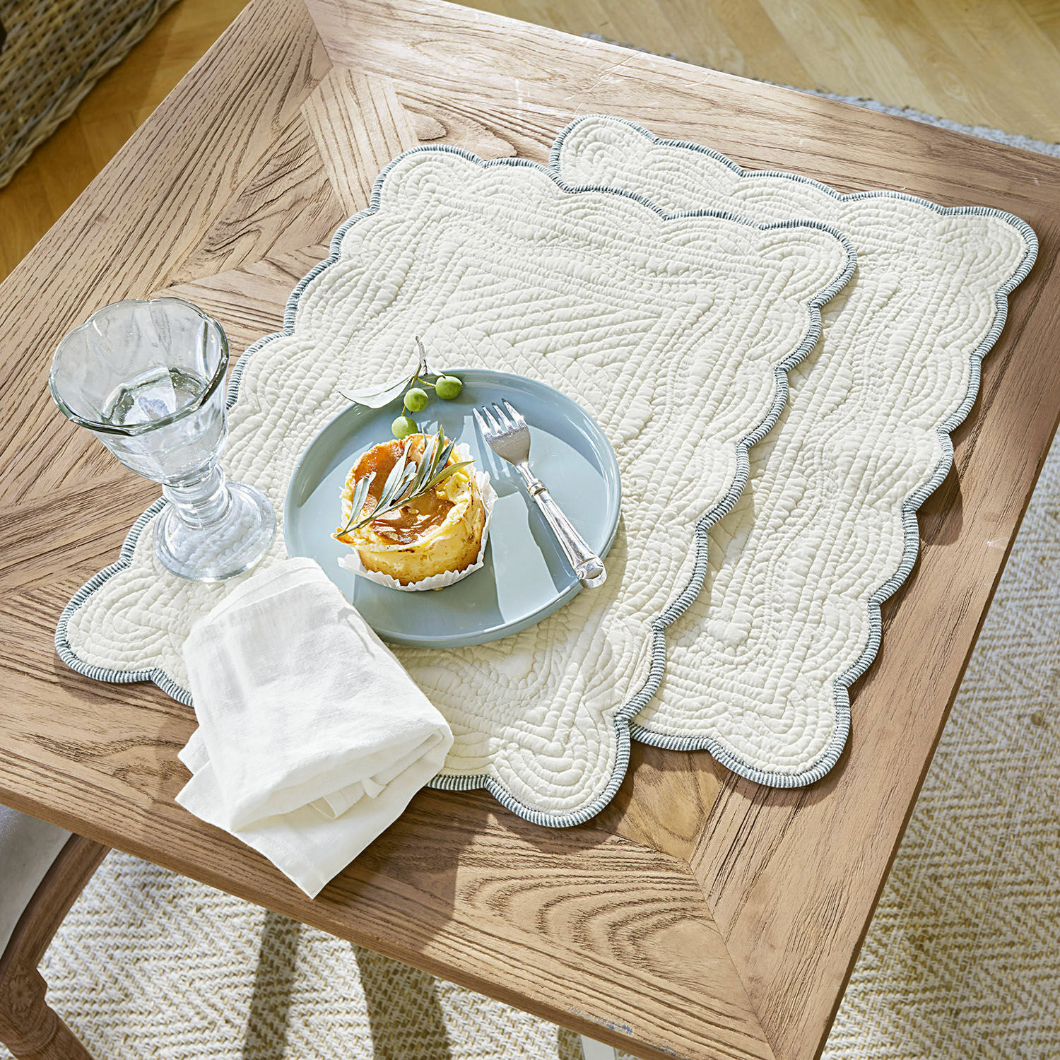 Platzset 2er Set Fontienne Beige gesteppte Baumwoll-Tischsets mit blauem Rand auf Holztisch mit Dessert und Glas