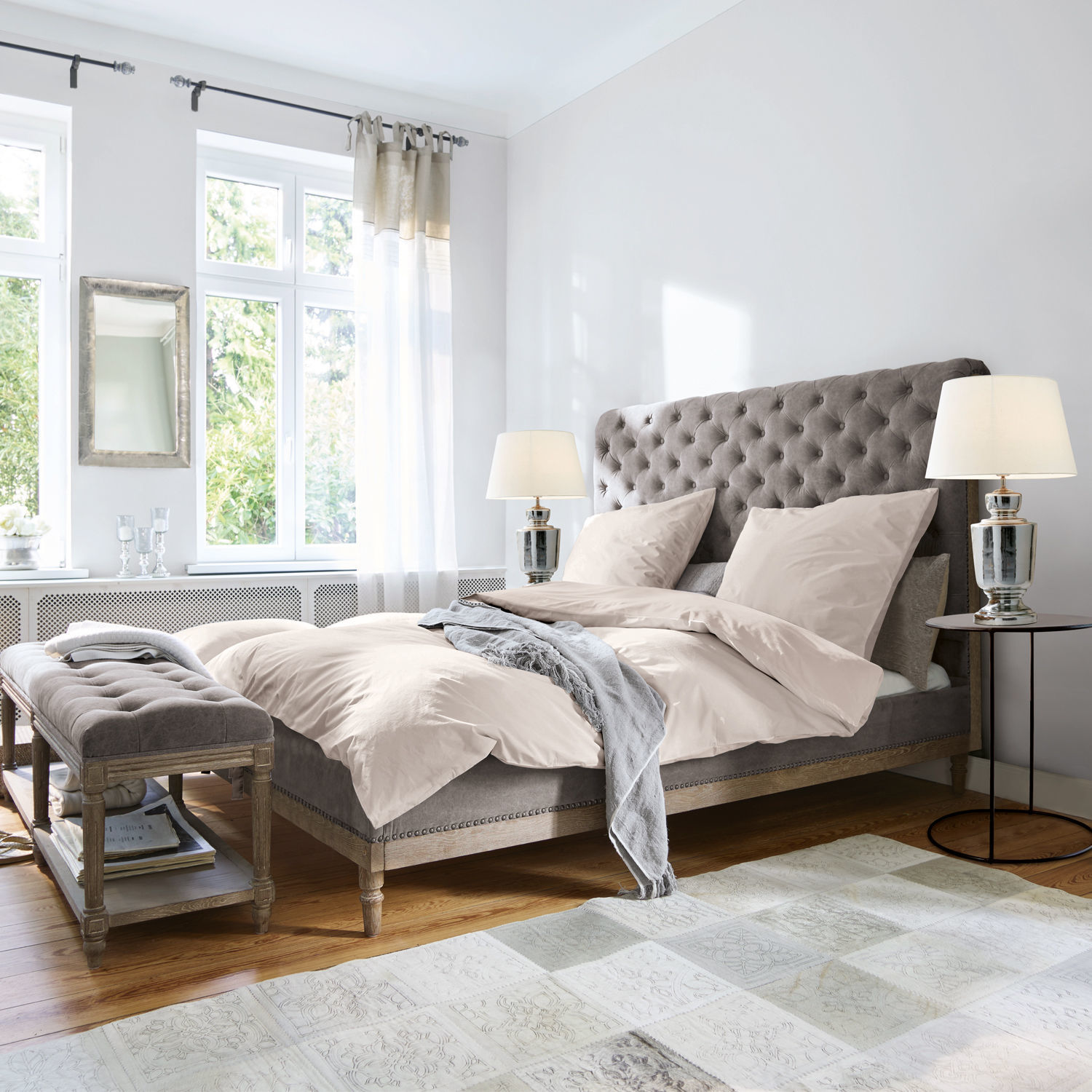 Bett Pozières Grau gepolstertes Bett mit gesteppter Kopfteil und beiger Bettwäsche in hellem Schlafzimmer mit Holzboden