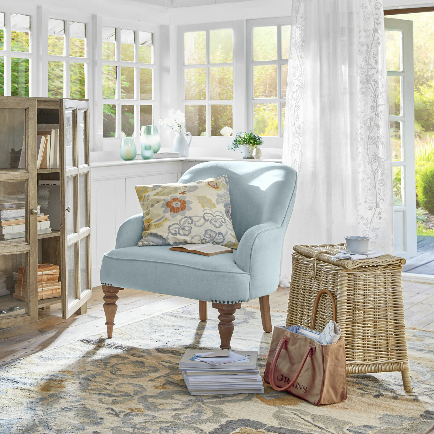 Sessel Ainsley Hellblauer Polsterstuhl mit Holzbeinen in hellem Wohnzimmer mit Korb aus Rattan und floralem Kissen