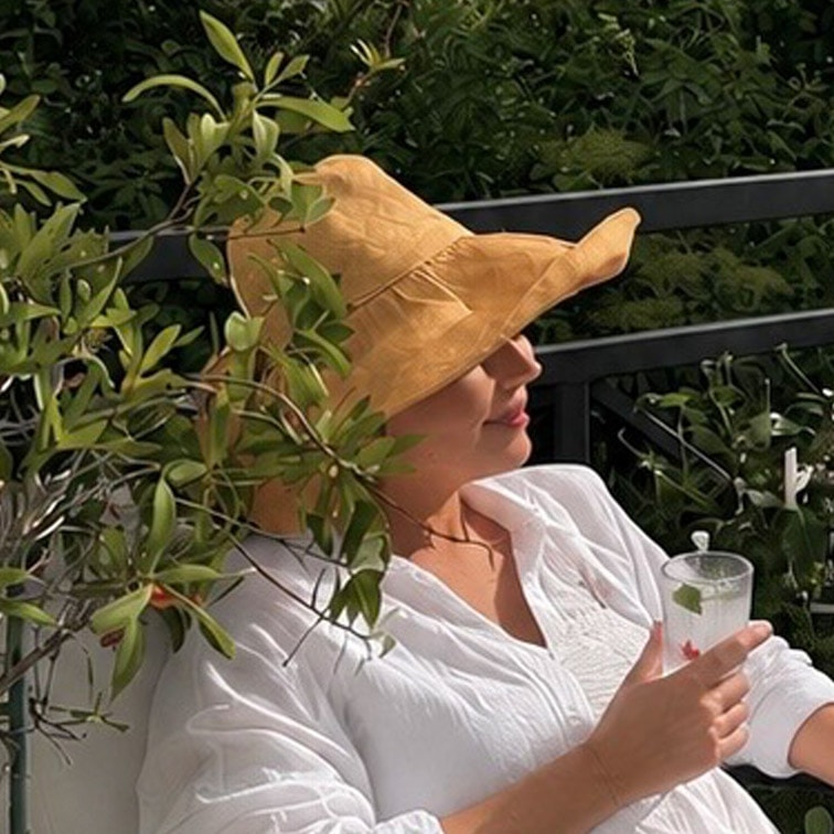 Frau in weißer Bluse und gelbem Strohhut entspannt auf Terrasse mit Getränk in der Hand