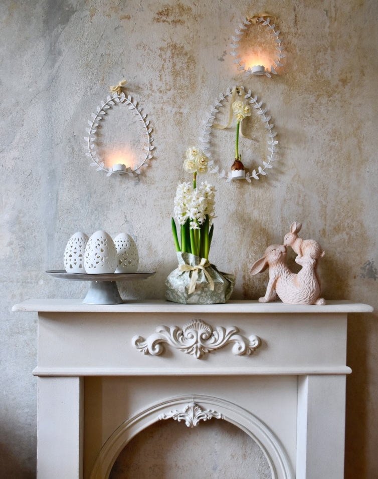 Pastellfarbene Osterdekoration mit weißen Keramikeiern, Blumen und Hasenfiguren auf einem Kaminsims