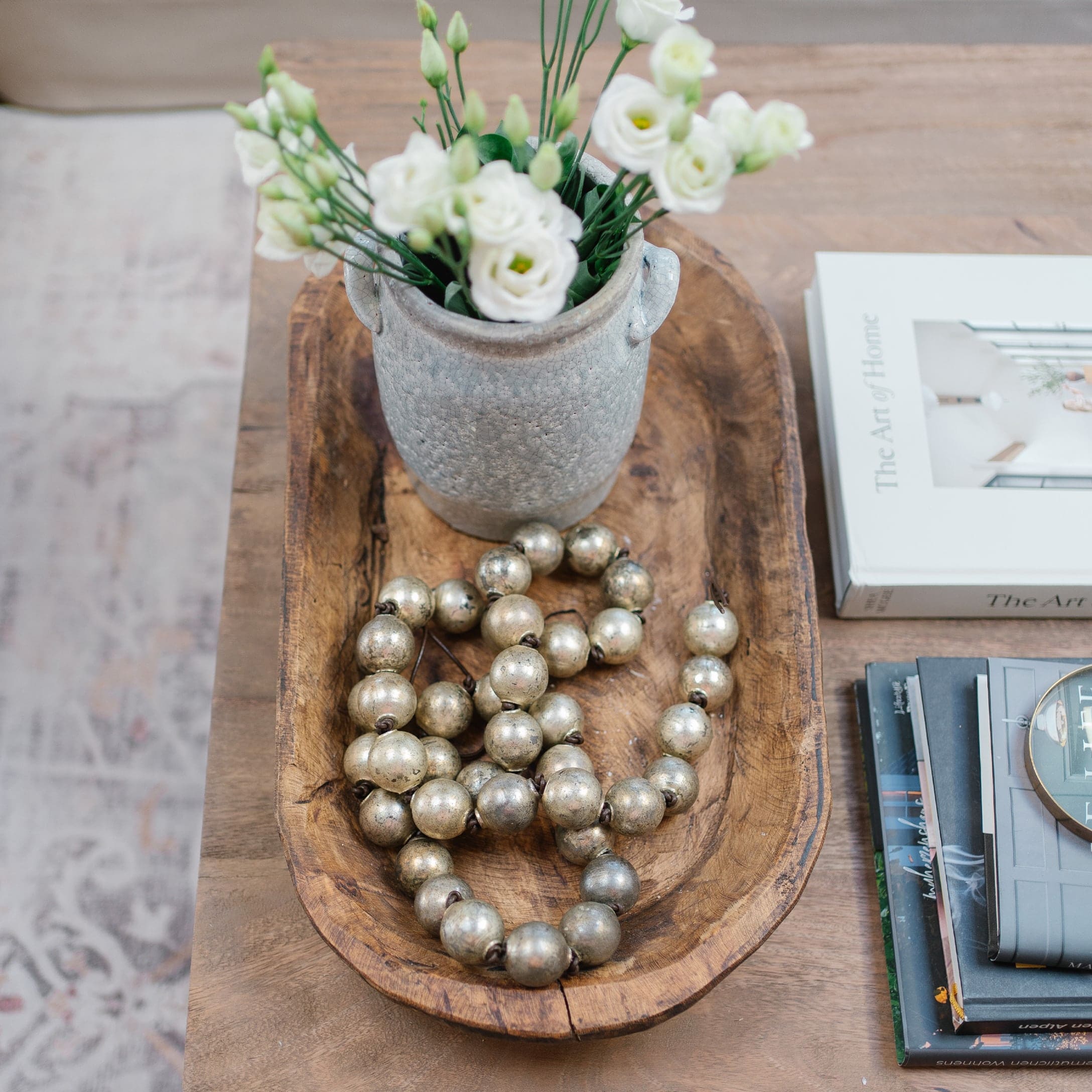Holzschale mit grauem Keramikvase, weißen Blumen und dekorativer Metallperlenkette auf Holztisch
