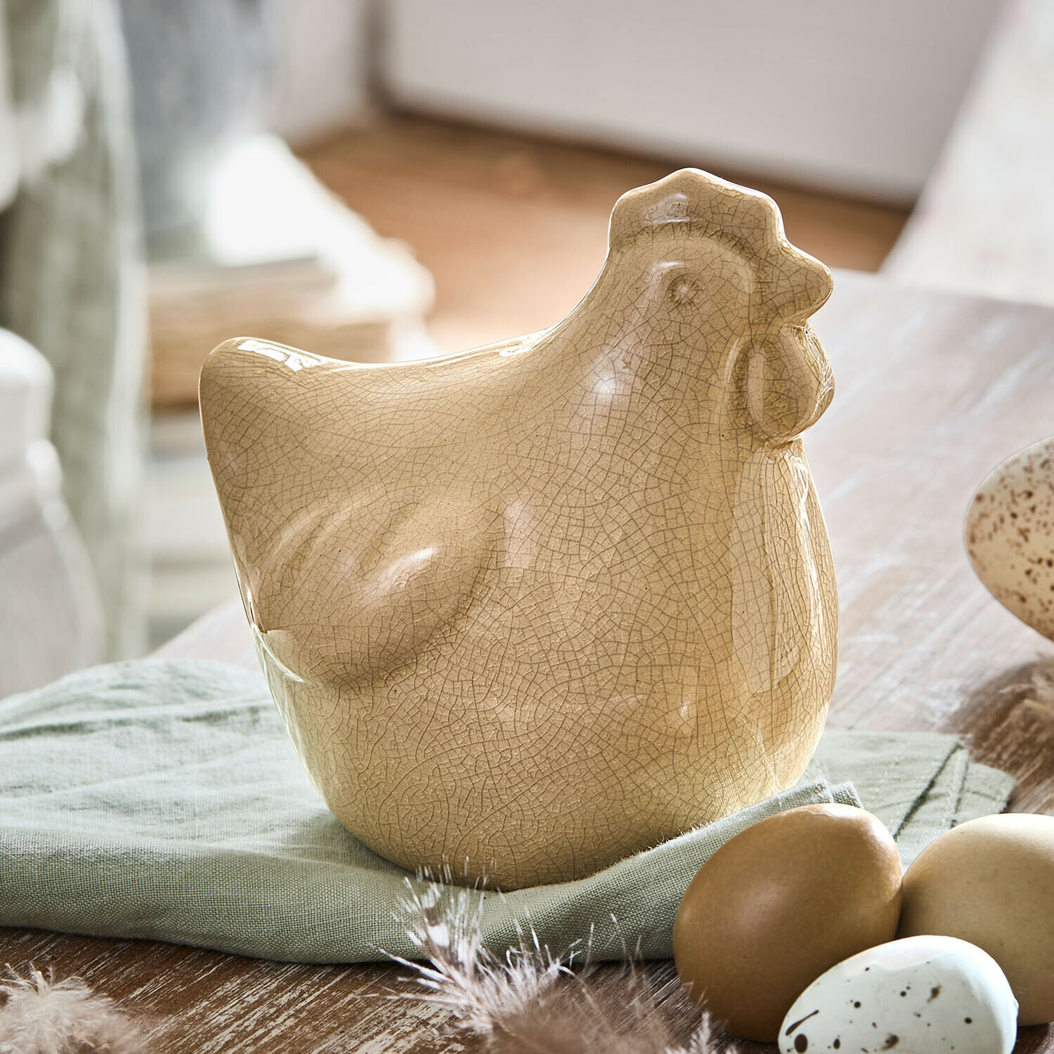 Beige Keramik-Hühnerfigur mit Craquelé-Effekt auf grünem Stoff neben dekorativen Eiern
