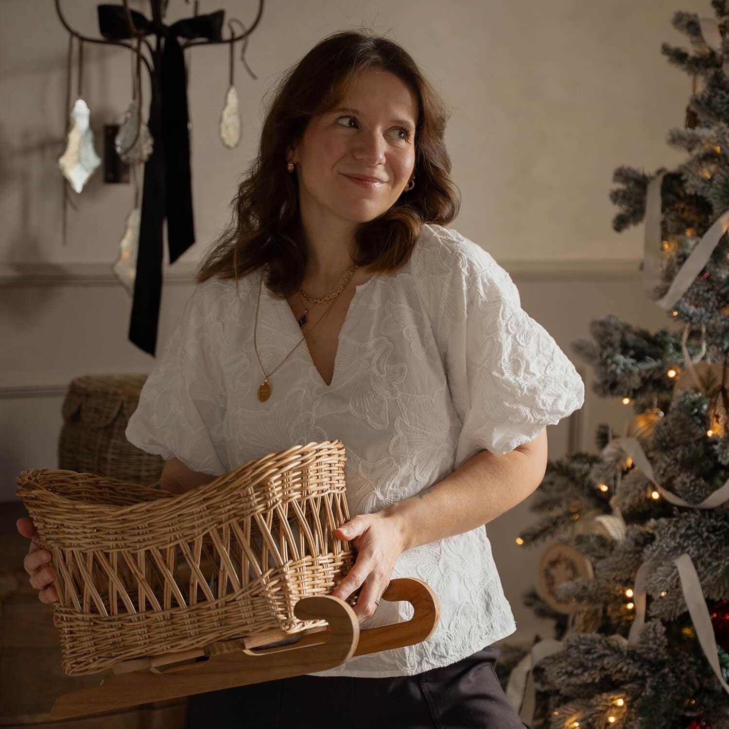 Frau in weißer Bluse hält einen Korbschlitten aus Weide mit Holzgestell neben geschmücktem Weihnachtsbaum