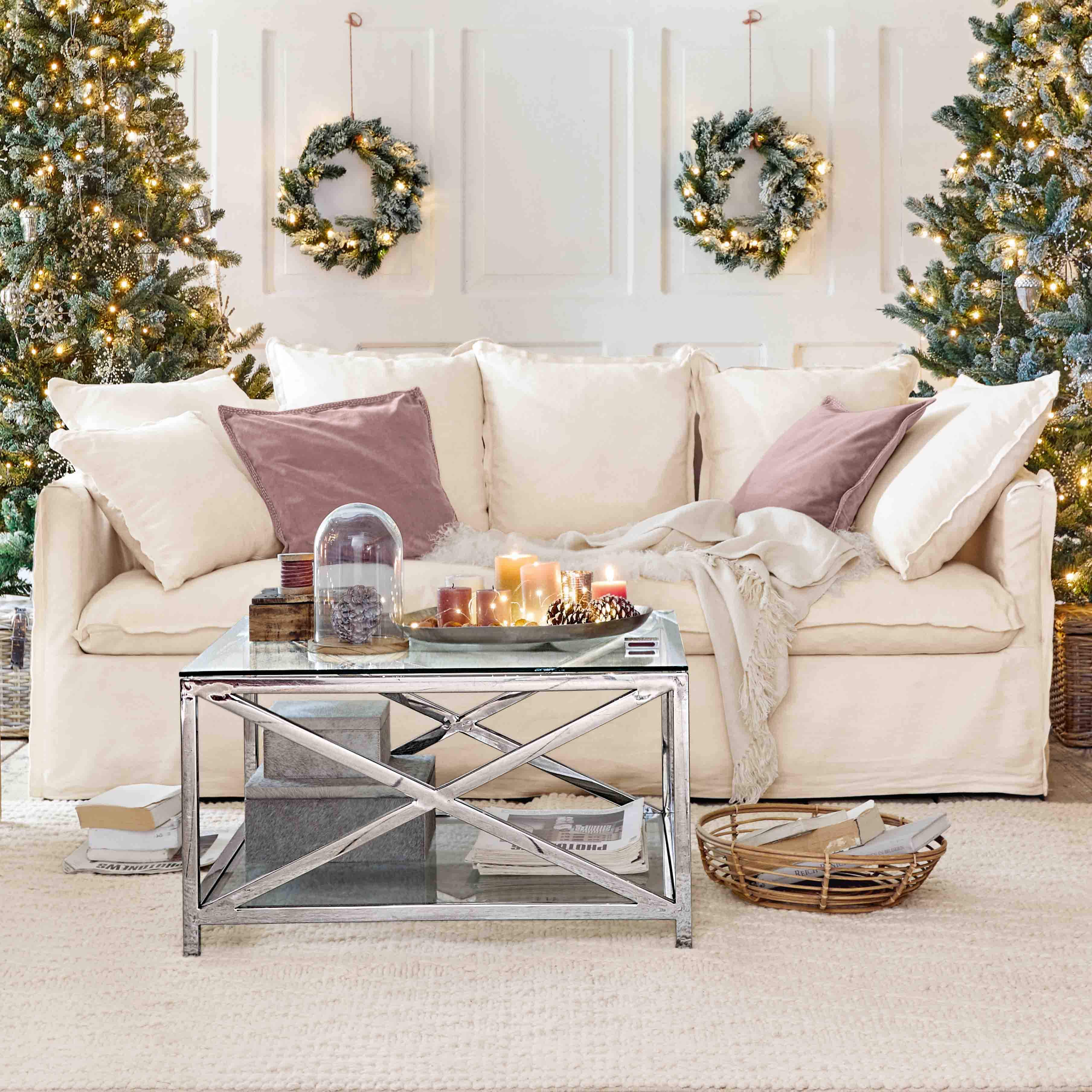 Bequeme cremefarbene Couch mit Kissen und Metall-Couchtisch vor weihnachtlicher Dekoration mit Tannenbäumen und Kränzen