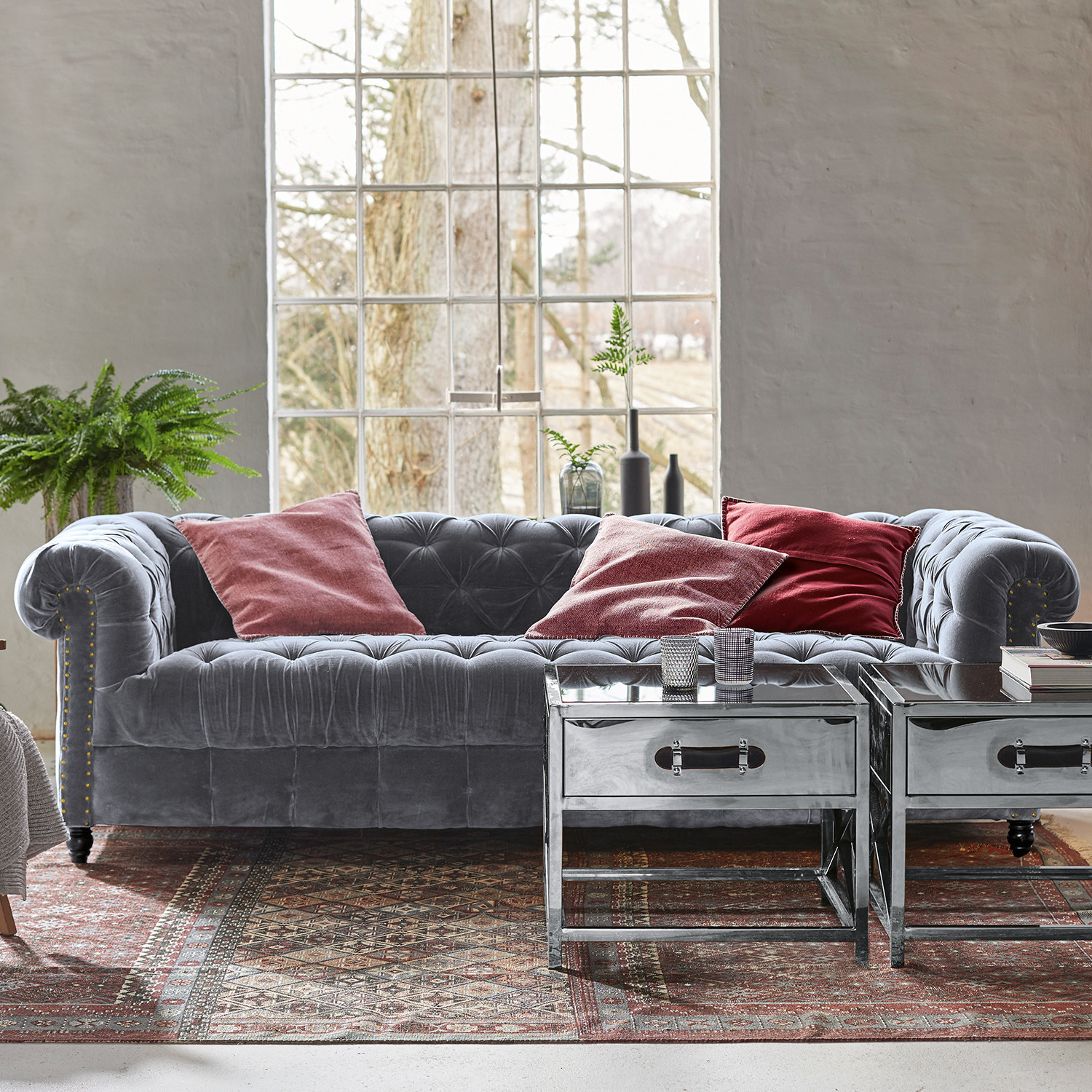 Sofa Parpeville Graues Samt-Chesterfield-Sofa mit roten Kissen und metallischen Beistelltischen auf gemustertem Teppich