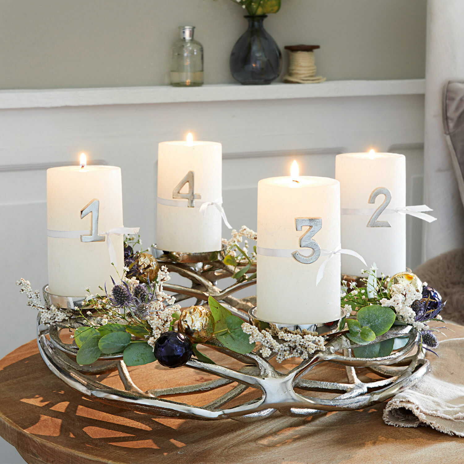 Adventskranz aus silbernem Metall mit vier weißen nummerierten Kerzen und grüner Dekoration