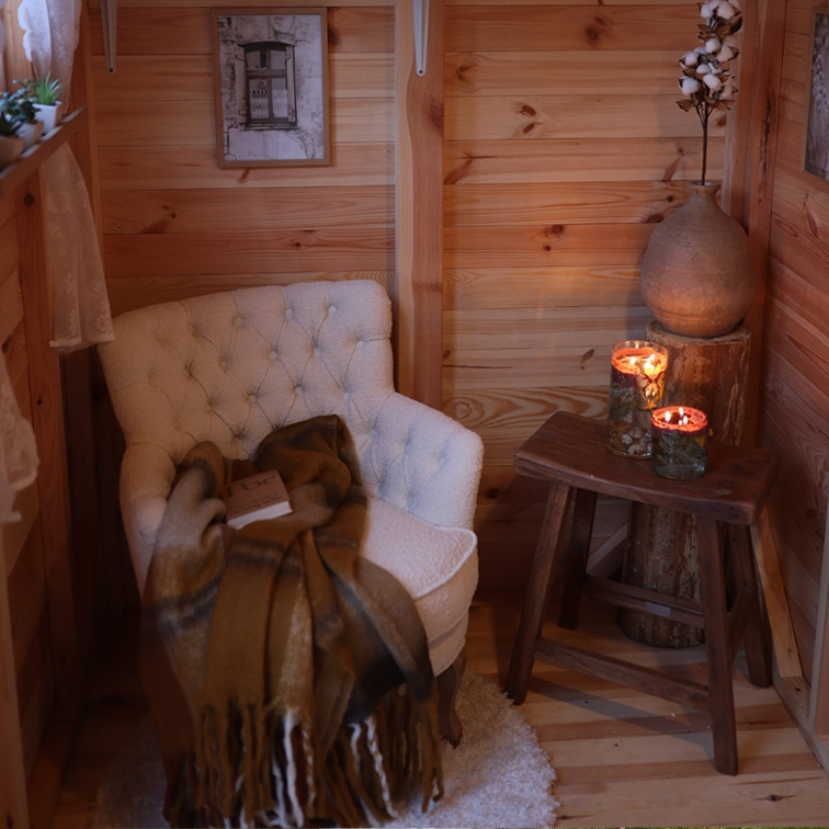 Gemütliche Ecke mit weißem, gepolstertem Sessel, braunem Plaid und Holzschemel mit brennenden Kerzen
