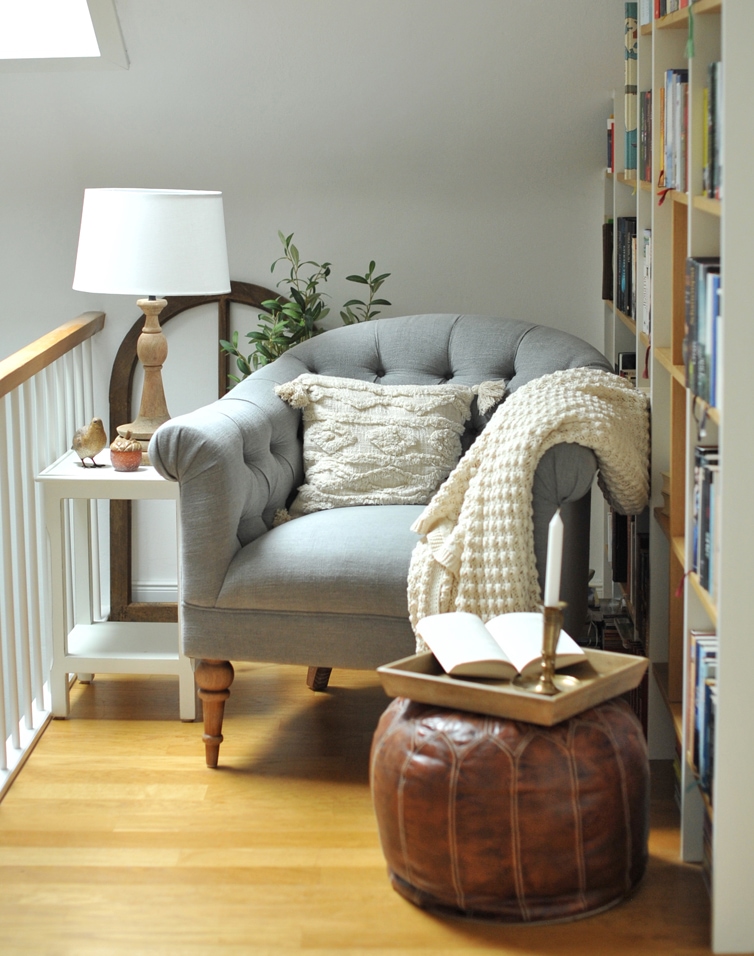 Grau gepolsterter Sessel mit Kissen und cremefarbener Decke neben braunem Lederhocker und Lampe