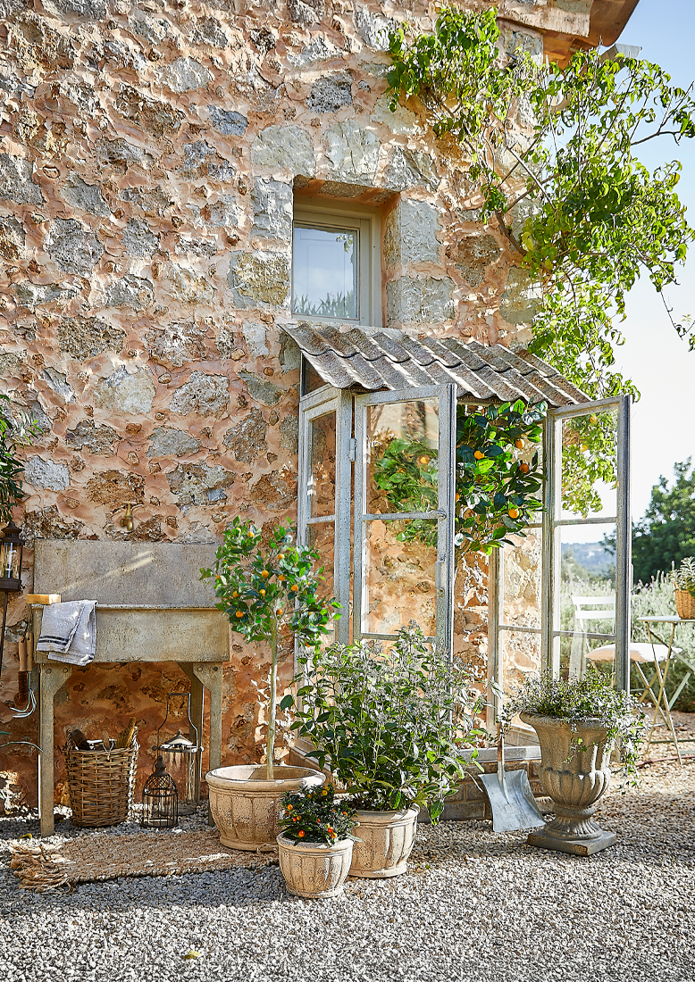 Garten mit Steinmauer, Metall-Gewächshaus und Pflanzgefäßen auf Kiesboden