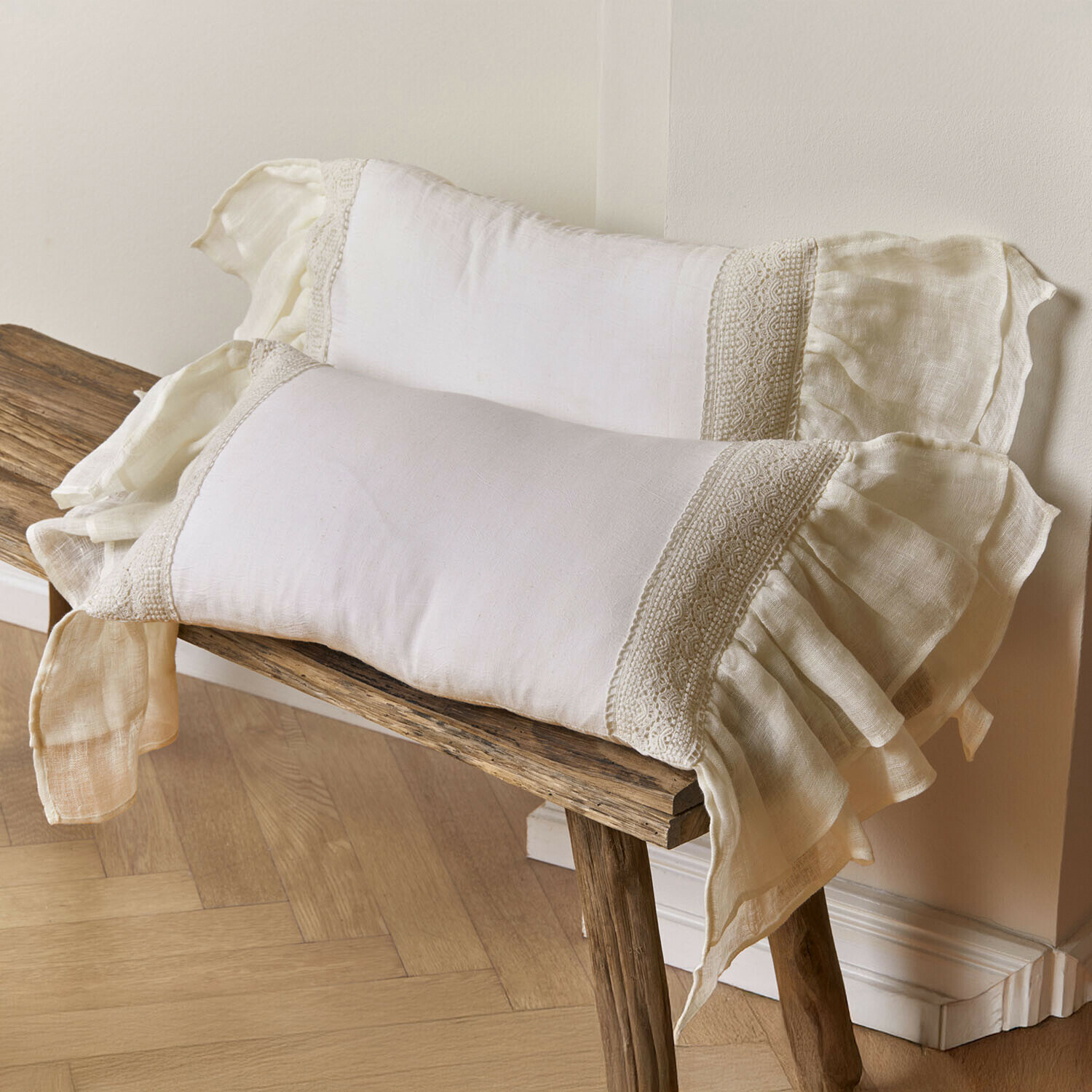 Kissenhülle 2er Set Jolévianne Zwei cremefarbene Leinenkissen mit Spitzenverzierung und Rüschen auf einer Holzbank