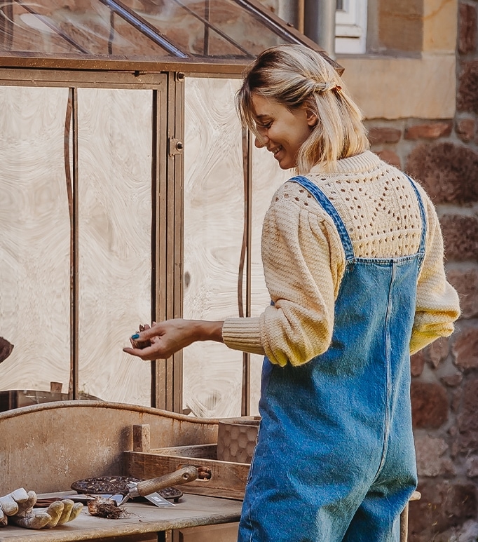Frau in blauem Latzhosen und cremefarbenem Pullover arbeitet an einem Holztisch im Garten
