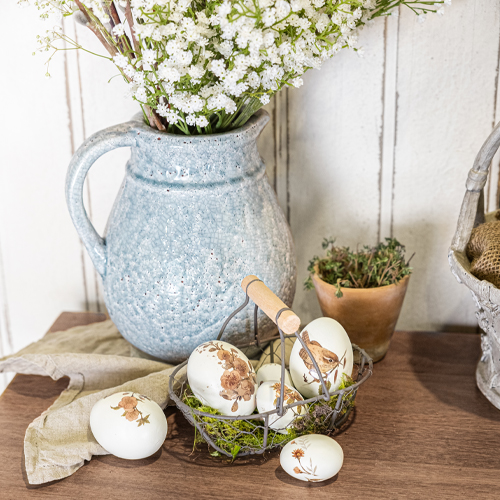 Blauer Keramikkrug mit weißen Blumen und dekorativen Eiern mit Tiermotiven auf Holztisch