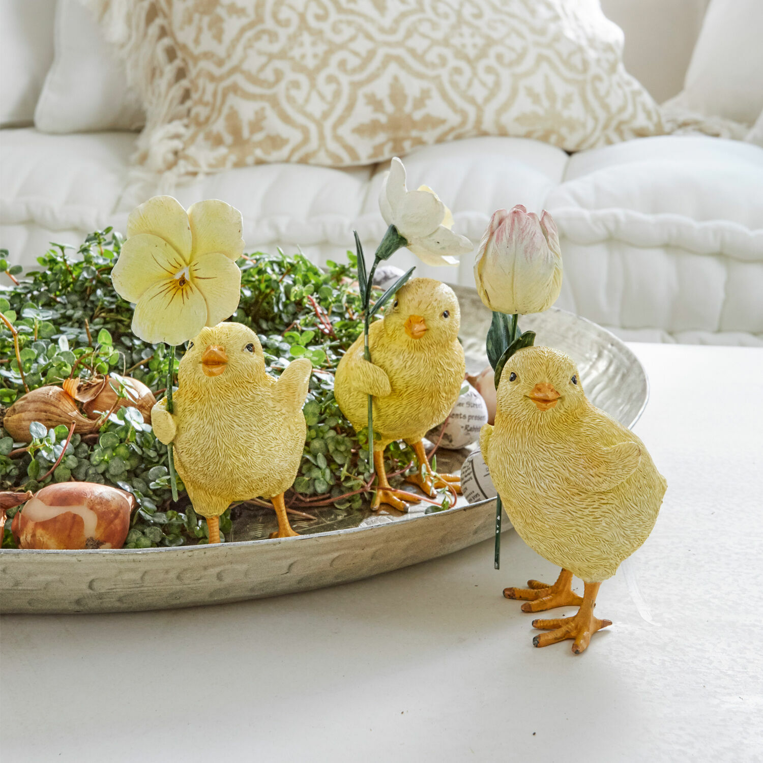 Gelbe Deko-Küken mit Blumen auf Metalltablett mit grünen Pflanzen, frühlingshafte Tischdekoration