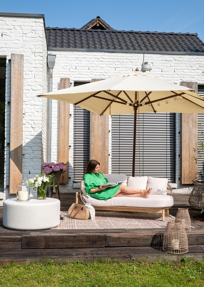 Frau im grünen Kleid entspannt auf hellem Sofa unter cremefarbenem Sonnenschirm auf Holzterrasse