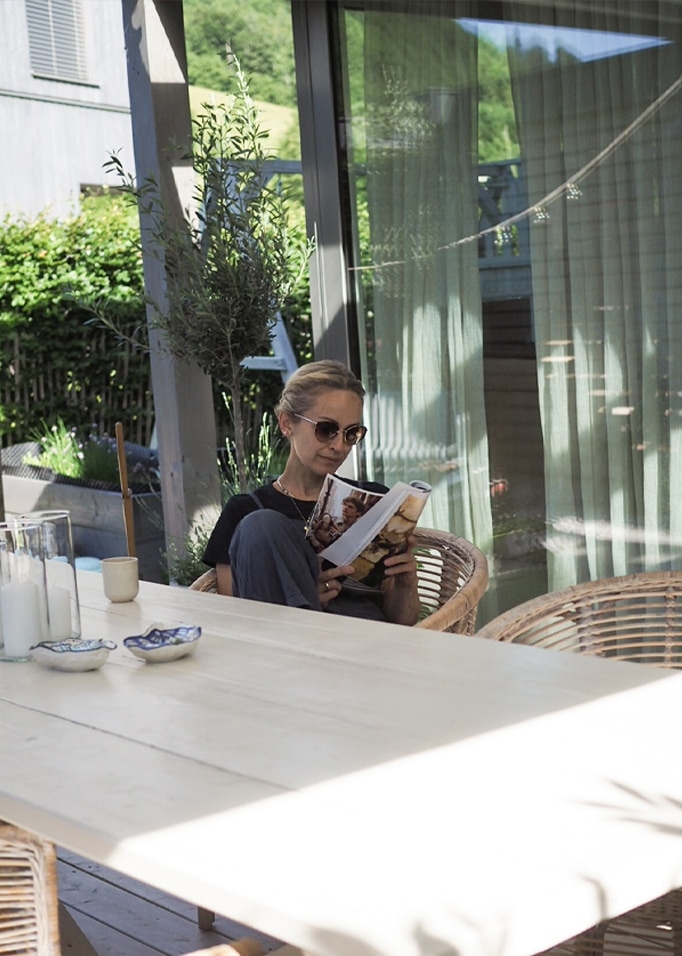 Frau liest Buch auf Terrasse mit hellem Holztisch und Rattanstühlen im Sonnenlicht